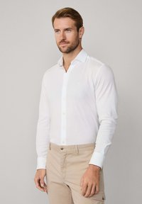 Biała koszula z długim rękawem wykonana z gładkiego materiału, z kołnierzykiem na guziki i jasnymi guzikami, połączona z beżowymi spodniami.