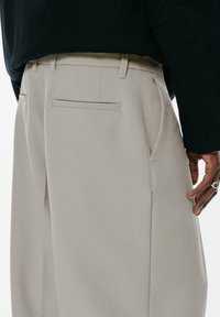 Beige Hose mit lockerer Passform, ausgestattet mit seitlichen Taschen und einer einzelnen Gesäßtasche. Glatte Textur und maßgeschneiderte Designdetails.