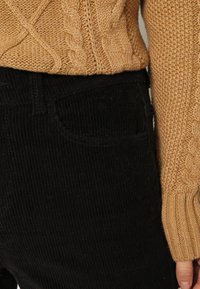 beige kabelstickad tröja i kombination med svarta kordflortbyxor med en framficka och vertikal ribbad textur. Avslappnat outfitdetalj.