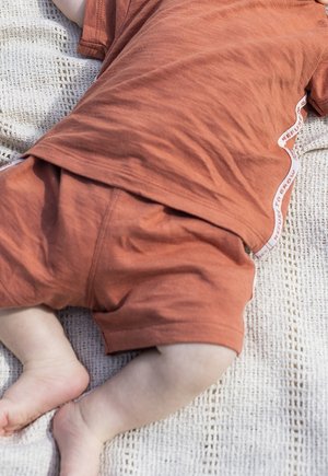 Bébé allongé sur une couverture tricotée portant un short couleur rouille et une chemise à manches courtes assortie avec une bordure blanche sur le côté.