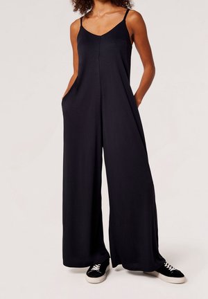 Kvinde iført en sort ærmeløs jumpsuit med vide ben og lommer samt sorte sneakers med hvide såler og snørebånd.