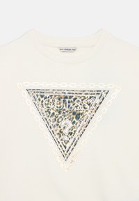 Jasno beżowy sweter z nadrukiem w kształcie trójkąta przedstawiającym lamparta z metalicznymi akcentami, szczegółowym tekstem i wzorem łańcucha wokół logo.