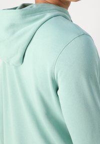 Mátově zelená mikina s kapucí s texturovaným povrchem. Má stahovací kapuci a žebrované manžety. Tkanina působí měkce a lehce.