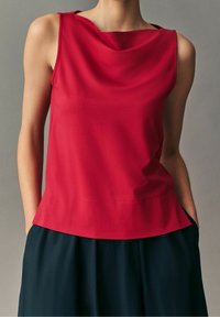 Haut sans manches rouge avec un col drapé, une texture lisse et un ourlet évasé. Il est associé à une jupe foncée, affichant une coupe décontractée.