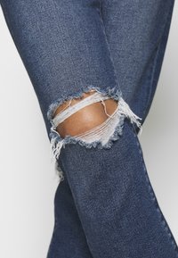 Blå denimjeans med fransade ripor på knäna, i en mörk tvätt och med en mjuk textur. Ripsen avslöjar ett ljusare inre tyg-detalj.