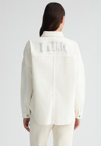 Giacca in denim bianco con vestibilità rilassata, caratterizzata da un logo "LIUJO" in strass argentati sul retro e polsini con bottoni con orlo sfilacciato.