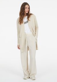 Beige Strickjacke mit offenem Front, gerippten Bündchen und zwei Taschen, kombiniert mit einem weißen T-Shirt und cremefarbenen weiten Hosen. Strukturierte Oberfläche.
