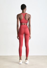 Vrouw staat met de rug naar ons toe in een rode Calvin Klein sportbeha en bijpassende leggings, met witte sneakers, op een effen achtergrond.