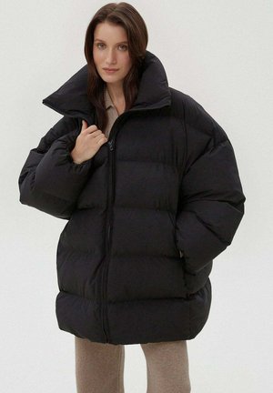 Zwarte pufferjack met een hoge kraag, gewatteerd ontwerp en oversized pasvorm. De ritssluiting en pluizige textuur verhogen de warmte en stijl.
