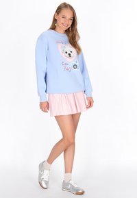 Világoskék pulóver kölyökkutya grafikával és felirattal, rózsaszín redős szoknyával és ezüst sportcipőkkel párosítva. Egyszerű fülbevalók teszik teljessé a megjelenést.
