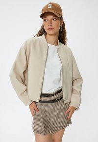 Chaqueta bomber beige, con cierre de cremallera, cuello de canalé, combinada con una camiseta blanca y una falda mini a cuadros con dos cinturones negros. Gorra marrón con logo.