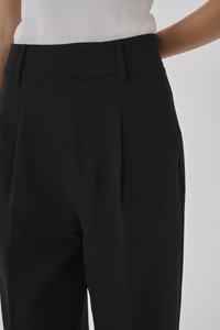 Svarta skräddarsydda byxor med hög midja, med frontveck och en slät textur, delvis synliga på bilden.