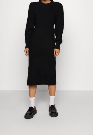 Person iført en sort midi-kjole med lange ærmer og puffede skuldre, hvide sokker og sorte loafers, der står mod en ensfarvet baggrund.