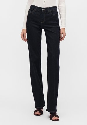 Femme portant un jean noir droit, un haut blanc à manches longues et des sandales ouvertes marron foncé, debout devant un fond uni.