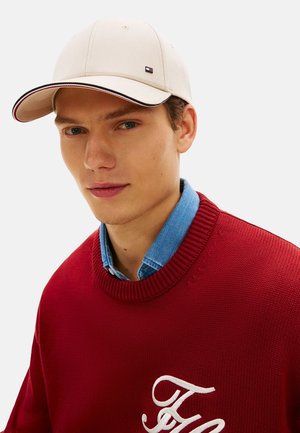 Jonge man met een beige pet, een blauw denim overhemd en een rode trui met wit geborduurde initialen op de borst, die vooruit kijkt.