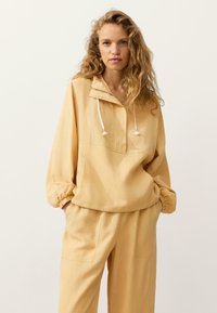 Femme aux cheveux bouclés portant un pull beige ample avec des cordons blancs et un pantalon large assorti, mains dans les poches, fond neutre.