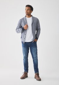 Gray knitted cardigan with a shawl collar, buttoned front, and navy accents. Worn over a white t-shirt and paired with blue jeans and brown boots.