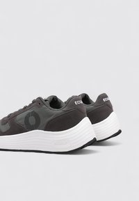 Zapatillas grises de Ecoalf con una parte superior lisa y texturizada, que presentan un logo circular prominente y suelas blancas en capas para mayor soporte.