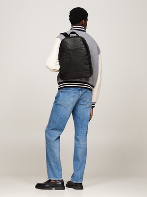 Schwarzer Rucksack mit gemustertem Design und glatter Textur, rechteckiger Form und einer vorderen Reißverschlusstasche, die auf dem Rücken einer Person in einer grauen Jacke und Jeans getragen wird.
