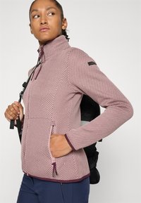 Ung kvinne iført en teksturert rosa zip-up-jakke med hånda i lommen og bærende en svart ryggsekk over skulderen.