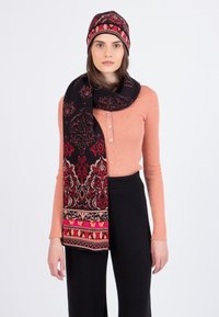 Sort uldne tørklæde med indviklede røde og lyserøde blomster mønstre, parret med en matchende strikket sort og rød hat. Modellen bærer en lys pink top.