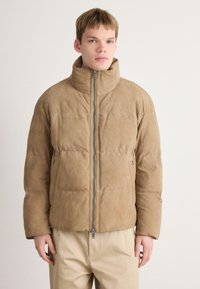 Brun ruskind pufferjakke med høj krave, frontlynlås og sidelommer. Har et quiltet design og en afslappet pasform.