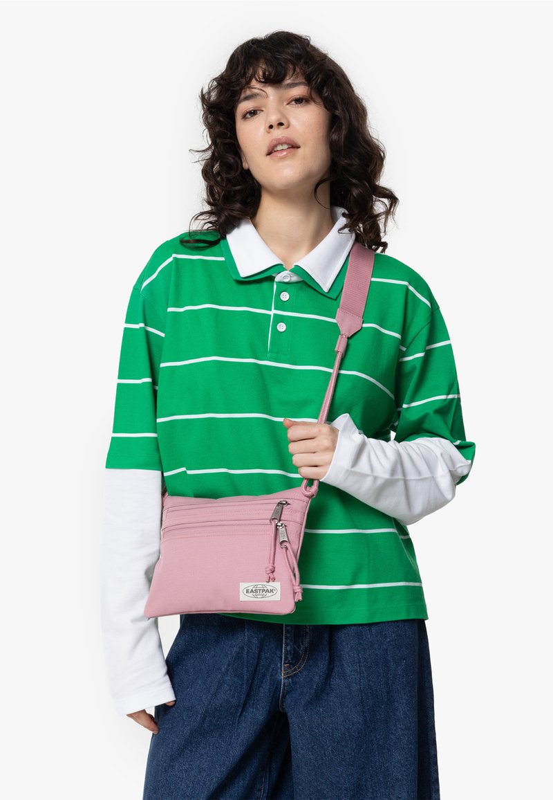Petit sac en bandoulière rose avec deux compartiments zippés, fabriqué en tissu doux. Présente une longue sangle réglable et une étiquette logo blanche.