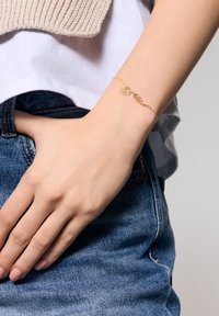 Złota bransoletka z misternym wzorem liści i kwiatów, z ogniwami łańcucha, noszona na nadgarstku ponad dżinsowym materiały, prezentująca codzienny strój.