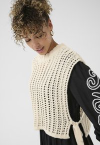 Top de tank cropped de punto en crema con patrón de puntada abierta, con un escote acanalado y lazos en los lados. Llevado sobre un vestido negro de manga larga.