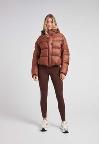 Brun pufferjakke med hætte, der har et quiltet design og knapper down fronten. Båret med brune leggins og hvide sneakers.