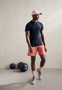 Svart Nike kortermet skjorte og rosa shorts; grå treningssko med detaljer; rosa caps; bakgrunnen inkluderer svarte treningsballer.