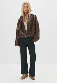 Giacca oversize in ecopelle marrone con cintura, abbinata a jeans a campana blu scuro e scarpe slip-on marroni con dettagli borchiati.
