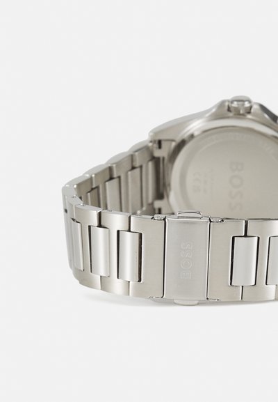 Montre en acier inoxydable argenté avec un bracelet à maillons texturé et un boîtier rond. Le fermoir est gravé du nom de la marque, BOSS.
