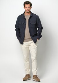 Dunkelblaue strukturierte Button-Up-Jacke mit zwei Brusttaschen, beigem T-Shirt, cremefarbenen Hosen und braunen Sneakers.
