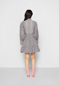 Žena s dlouhými tmavými vlasy, oblečená v květinových šatech s krajkovými detaily a růžovými kotníkovými botami na podpatku, stojící zády k jednoduchému pozadí.