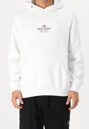 Homme portant un sweat à capuche blanc Tommy Hilfiger avec poche avant et un pantalon noir.