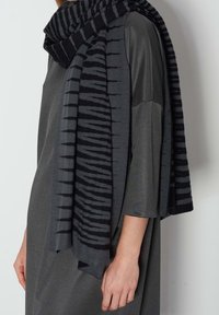 Femme portant une robe gris foncé à manches trois-quarts et une écharpe rayée noire et grise drapée sur les épaules.