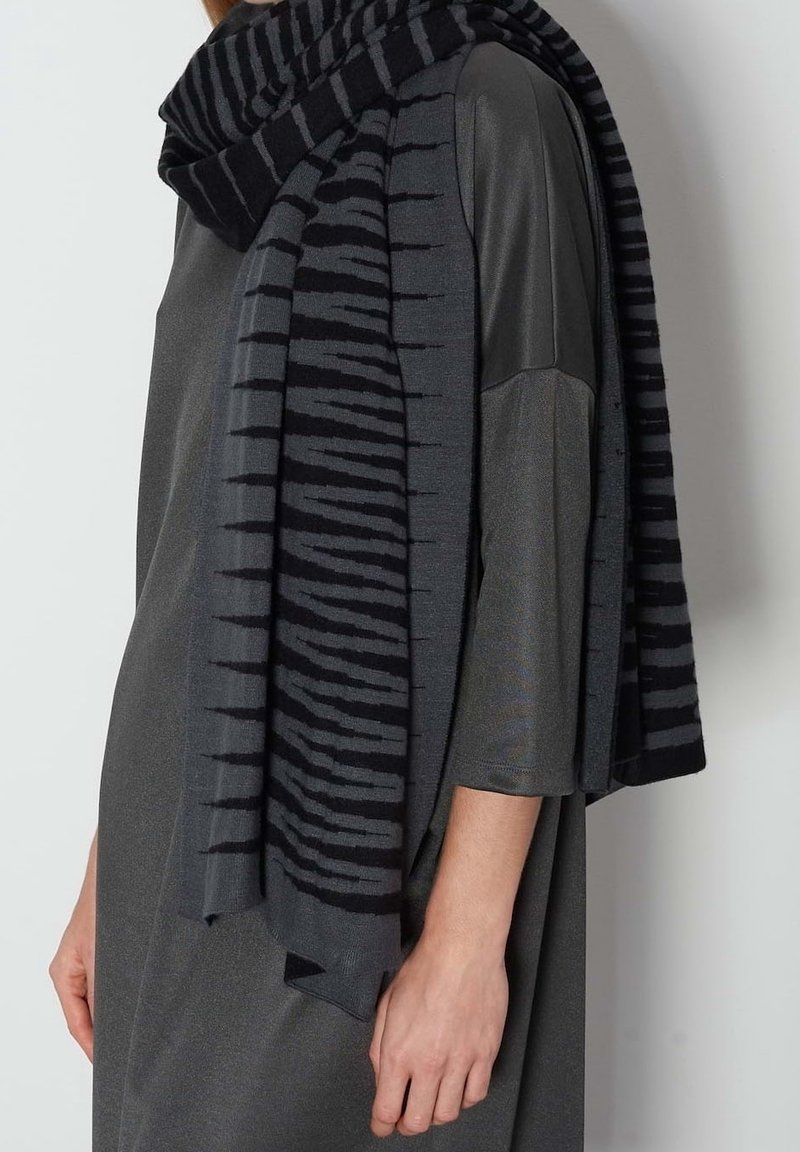 Femme portant une robe gris foncé à manches trois-quarts et une écharpe rayée noire et grise drapée sur les épaules.