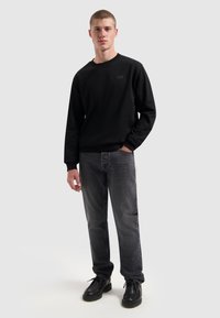 Zwarte sweatshirt met ronde hals en subtiele logo, gecombineerd met donkergrijze jeans en zwarte veterschoenen. Glad materiaal met een relaxed fit.