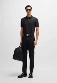 Black polo shirt, black trousers, black belt with silver buckle, and black bag. Glossy black shoes. Model wearing dark sunglasses.