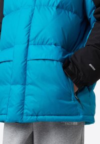 Puffig turkos jacka med svarta ärmar, med hög krage, sidofickor och elastiska muddar. Tillverkad av syntetiska material.