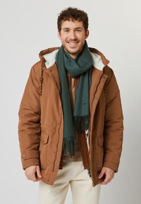 Parka marrón, aislante, con capucha forrada de borrego y bolsillos, combinada con una bufanda verde. Modelo vistiendo una blusa beige y pantalones claros.