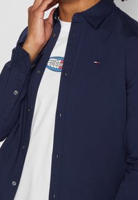 Marinblå skjorta med knappar och spetsig krage, prydd med en liten logotyp på bröstet. Under den en vit t-shirt med en färgglad grafisk design.