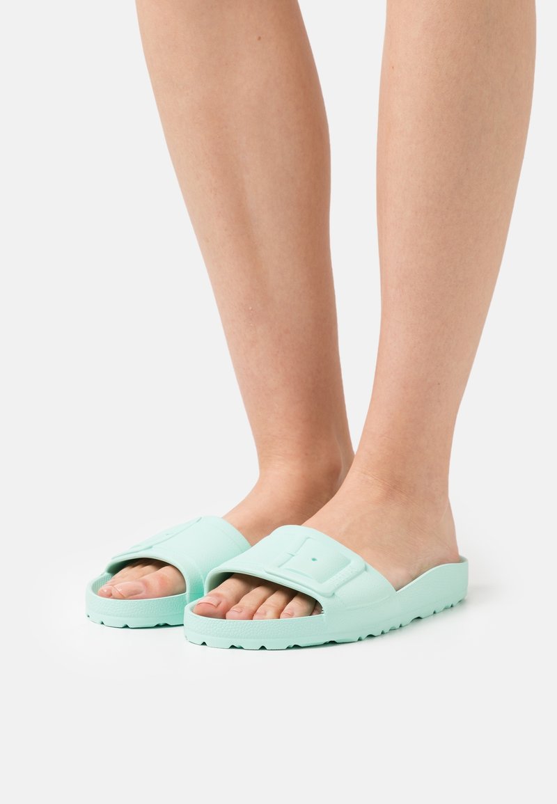 Pieds portant des sandales slides turquoise clair avec de larges brides ajustables et des semelles épaisses et texturées sur fond blanc.