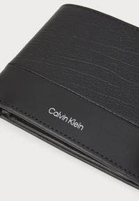 Svart lærpung med strukturert topp og glatt bunnseksjon. Inkluderer preget "Calvin Klein"-logo i hvitt.