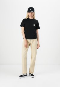 Black cotton t-shirt with a small white logo pocket, paired with beige straight-leg pants and black sneakers. Black cap completes the look.