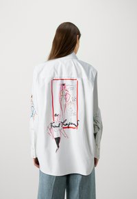 Biała koszula z długim rękawem, z kolorowymi szkicami i projektami mody nadrukowanymi na plecach, obramowanymi na czerwono, z dokładnymi ilustracjami.