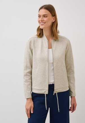 Leende kvinna klädd i en ljusgrå zip-up jacka över en vit topp, ihop med mörkblå byxor, mot en neutral bakgrund.