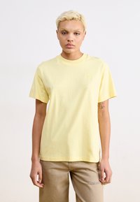 T-shirt en coton jaune clair à manches courtes et col rond. Il présente un petit logo brodé sur la poitrine et une coupe décontractée.