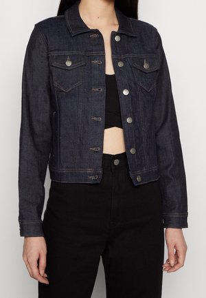 Femme portant une veste en jean bleu foncé avec des boutons argentés, un crop top noir et un pantalon noir taille haute sur fond uni.